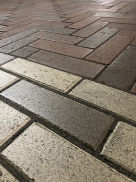 A close-up view of a brick pavement with a herringbone pattern. The bricks vary in color, including shades of gray, beige, and brown, creating a textured and ordered appearance.