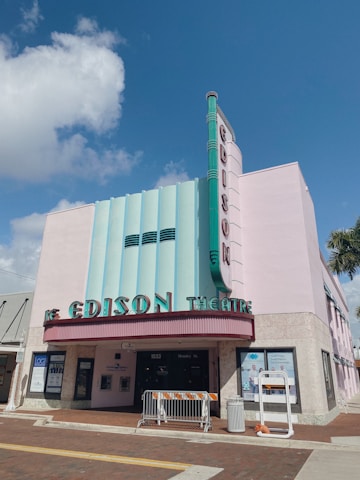 An Art Deco-style theater building with the name 'Edison Theatre' prominently displayed in bold letters. The facade features pastel pink and light blue colors with vertical lines and ornamental designs typical of the Art Deco period. A marquee indicates the entrance, and there are posters in the windows. The sky is clear and blue, complementing the building’s colors.
