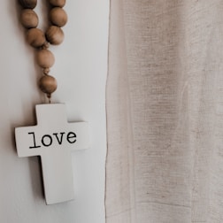 A white cross with the word 'love' written on it hangs on a wall. Next to it, a strand of wooden beads drapes downward. To the right, there is a sheer, textured curtain allowing soft light to pass through. The overall setting has a serene and peaceful atmosphere.