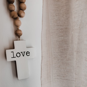 A white cross with the word 'love' written on it hangs on a wall. Next to it, a strand of wooden beads drapes downward. To the right, there is a sheer, textured curtain allowing soft light to pass through. The overall setting has a serene and peaceful atmosphere.