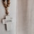 A white cross with the word 'love' written on it hangs on a wall. Next to it, a strand of wooden beads drapes downward. To the right, there is a sheer, textured curtain allowing soft light to pass through. The overall setting has a serene and peaceful atmosphere.