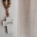 A white cross with the word 'love' written on it hangs on a wall. Next to it, a strand of wooden beads drapes downward. To the right, there is a sheer, textured curtain allowing soft light to pass through. The overall setting has a serene and peaceful atmosphere.