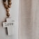 A white cross with the word 'love' written on it hangs on a wall. Next to it, a strand of wooden beads drapes downward. To the right, there is a sheer, textured curtain allowing soft light to pass through. The overall setting has a serene and peaceful atmosphere.