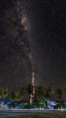 A starry night sky featuring the Milky Way galaxy stretches above a tall communication tower surrounded by palm trees. The ground is illuminated with artificial light, and a blur of motion suggests passing vehicles.
