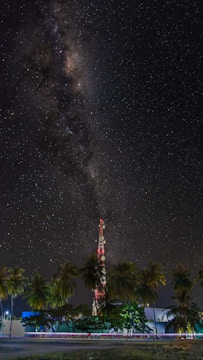 A starry night sky featuring the Milky Way galaxy stretches above a tall communication tower surrounded by palm trees. The ground is illuminated with artificial light, and a blur of motion suggests passing vehicles.