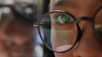 A close-up view of a person's eye, partially obscured by round glasses. The glasses reflect some green and ambient lighting. The background is out of focus, giving prominence to the eye and the reflection in the glasses.