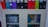 A shelf contains a row of colorful binders in shades of orange, blue, purple, black, red, and green on top. Below, a cabinet with glass doors displays products including boxes labeled 'Baby Cam' and hygiene items, along with a blue hand sanitizer bottle.