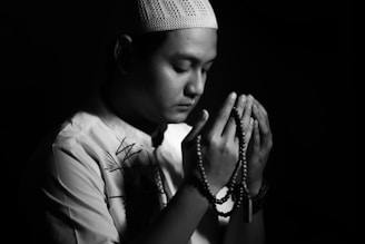 A person is sitting in a dark setting, holding prayer beads. The individual appears to be in a meditative or reflective state, with eyes closed, and is wearing a decorative shirt and a knit cap.