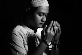 A person is sitting in a dark setting, holding prayer beads. The individual appears to be in a meditative or reflective state, with eyes closed, and is wearing a decorative shirt and a knit cap.