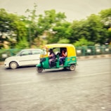 A green and yellow auto-rickshaw is driving alongside a white car on a road. The vehicle motion is captured with a blur effect, indicating speed. The background features green trees and a metal fence, suggesting an urban setting.