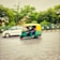 A green and yellow auto-rickshaw is driving alongside a white car on a road. The vehicle motion is captured with a blur effect, indicating speed. The background features green trees and a metal fence, suggesting an urban setting.