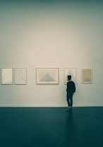 A person is standing in front of a gallery wall with five framed minimalist artworks, each featuring geometric shapes, primarily triangles. The setting appears to be a quiet, indoor exhibition space with a calm atmosphere.