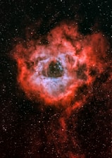 A vivid and detailed view of a nebula with a central bright region surrounded by swirling red clouds of gas and dust. The black background is dotted with numerous small stars scattered throughout the scene.