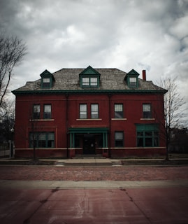 A historical red brick building with three dormer windows on the roof and a centered green front door, set against a cloudy sky. The building has symmetrical features and trees are visible on either side.