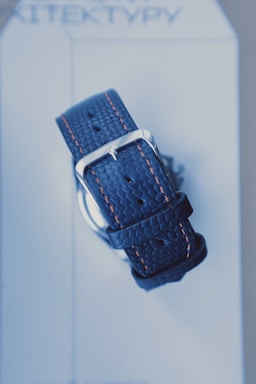 A close-up view of a textured blue watch strap with orange stitching and a silver buckle, placed on a light-colored background with text that appears partially visible.
