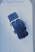 A close-up view of a textured blue watch strap with orange stitching and a silver buckle, placed on a light-colored background with text that appears partially visible.