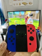 A person holds a Nintendo Switch controller with blue and red Joy-Cons in front of a TV displaying a colorful animated game scene featuring characters and a wooden fence. The room has a casual, homey atmosphere, with a small red flower vase visible on the side.