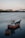 A small fishing boat with the name 'Ana Celia Vila do Conde' is moored in calm waters near a coastline. A smaller vessel is attached to it. In the background, industrial buildings and a crane are visible, suggesting a shipyard or port area.