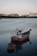 A small fishing boat with the name 'Ana Celia Vila do Conde' is moored in calm waters near a coastline. A smaller vessel is attached to it. In the background, industrial buildings and a crane are visible, suggesting a shipyard or port area.