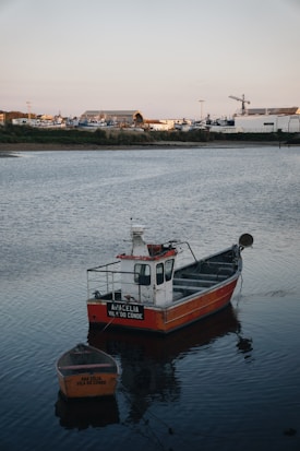 A small fishing boat with the name 'Ana Celia Vila do Conde' is moored in calm waters near a coastline. A smaller vessel is attached to it. In the background, industrial buildings and a crane are visible, suggesting a shipyard or port area.