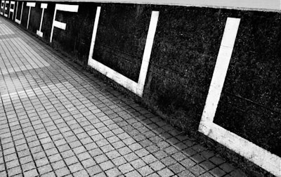 A black and white photograph depicting a concrete wall on which the word 'BEAUTIFUL' is painted in large, bold white letters. The wall is adjacent to a tiled pathway that features alternating light and dark tiles creating a textured, geometric pattern.