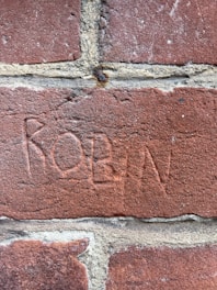 A close-up view of a red brick wall with the name 'ROBIN' etched into the surface of one brick. The grooves of the etching are visible against the rough texture of the brick. The mortar lines dividing the bricks are prominent and uneven.