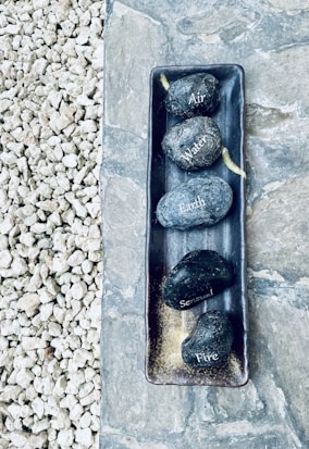 A collection of five smooth stones placed in a rectangular tray, each labeled with different words: Air, Water, Earth, Sensual, and Fire. The tray is positioned on a stone slab, adjacent to a section of small, loose gravel.