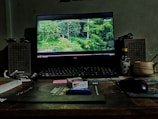 A desktop setup features a monitor displaying a scene with green trees on the screen. Two speakers flank the monitor. Various items, including a pack of Derby cigarettes, a lighter, a bottle, a cup, sunglasses, and a keyboard, are arranged on the wooden desk. The environment has a cozy, slightly cluttered look with muted lighting.