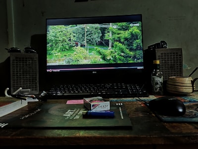 A desktop setup features a monitor displaying a scene with green trees on the screen. Two speakers flank the monitor. Various items, including a pack of Derby cigarettes, a lighter, a bottle, a cup, sunglasses, and a keyboard, are arranged on the wooden desk. The environment has a cozy, slightly cluttered look with muted lighting.
