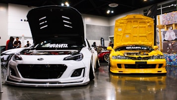Two cars are displayed indoors with their hoods open, showcasing their engines. The car on the left is white, and the car on the right is yellow. The scene includes several people in the background, along with other cars partially visible and various decorations on the walls.