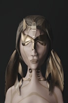 A stylized female figure with smooth, light-colored skin and a neutral expression wears an intricate gold and bronze mask that covers the eyes and nose, featuring geometric patterns. The figure has long, flowing hair adorned with large, metallic, ribbon-like decorations. The neck and upper chest have ornate metallic ornaments with similar geometric designs.