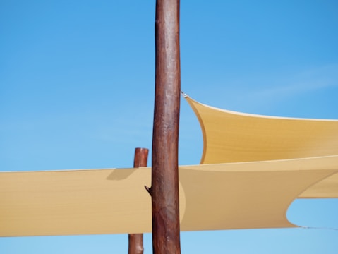 Two wooden poles are visible against a clear blue sky, with yellow shade sails attached to them. The sails are tightly stretched, creating geometric shapes against the sky.