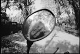 A black and white photograph captures a scene reflected in a round motorcycle mirror, showing part of a person's face and camera. The background consists of a tree-lined street with dappled sunlight filtering through the leaves, creating a contrast between shadows and light.
