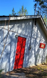 A small, rustic building with white wooden siding and a bright red door. Shadows from nearby trees create an intricate pattern on the wall. A small red-framed window is visible, and leafy trees surround the building under a clear blue sky.