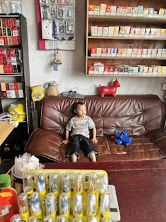 A young child sits on a brown leather sofa in a room that seems to be a small shop or convenience store. Behind the child, there are shelves filled with various cigarette packs. There is a red toy horse on the shelf. To the left, there's a black shelving unit with more packed items and display merchandise. A table in front of the sofa has a variety of small items in yellow packaging, possibly lighters or batteries.