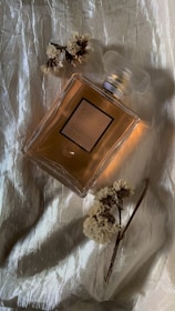 A perfume bottle with a label 'Coco Chanel' is placed on a textured, shiny fabric background. Beside the bottle, dried flowers are artfully arranged, adding an elegant touch to the composition.