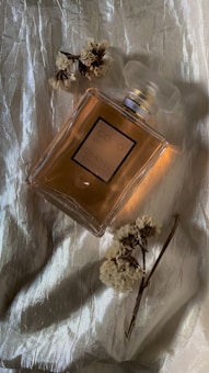 A perfume bottle with a label 'Coco Chanel' is placed on a textured, shiny fabric background. Beside the bottle, dried flowers are artfully arranged, adding an elegant touch to the composition.