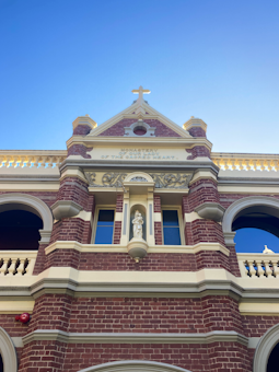 A red-brick building with cream-colored details, featuring arched windows and decorative stonework. The central part of the building has a prominent cross at the top, with a statue enshrined below it. Above the statue, there's an inscribed text that reads 'Monastery of Our Lady of the Sacred Heart'. The structure is well-lit, set against a clear blue sky.