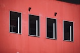 Four rectangular windows line a vibrant red wall. The windows have dark glass and white frames. Two small black square fixtures are mounted above the windows.