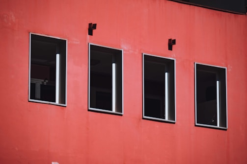Four rectangular windows line a vibrant red wall. The windows have dark glass and white frames. Two small black square fixtures are mounted above the windows.