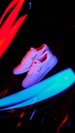 A pair of white sneakers with a prominent logo on the side is placed against a dark background. Bright neon streaks of red and blue light surround the shoes, creating a vibrant and energetic atmosphere.