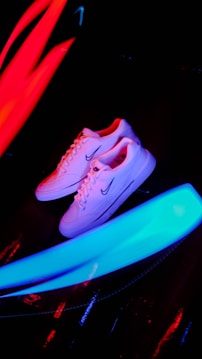 A pair of white sneakers with a prominent logo on the side is placed against a dark background. Bright neon streaks of red and blue light surround the shoes, creating a vibrant and energetic atmosphere.