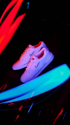 A pair of white sneakers with a prominent logo on the side is placed against a dark background. Bright neon streaks of red and blue light surround the shoes, creating a vibrant and energetic atmosphere.