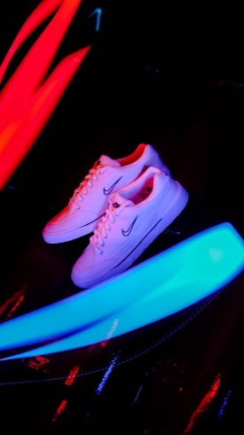 A pair of white sneakers with a prominent logo on the side is placed against a dark background. Bright neon streaks of red and blue light surround the shoes, creating a vibrant and energetic atmosphere.