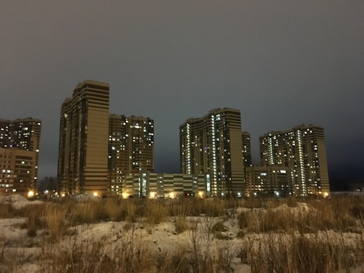A group of tall, modern apartment buildings illuminated with artificial lights against a cloudy night sky. Sparse grasses and patches of snow cover the ground in the foreground, creating a contrast between urban and natural elements.