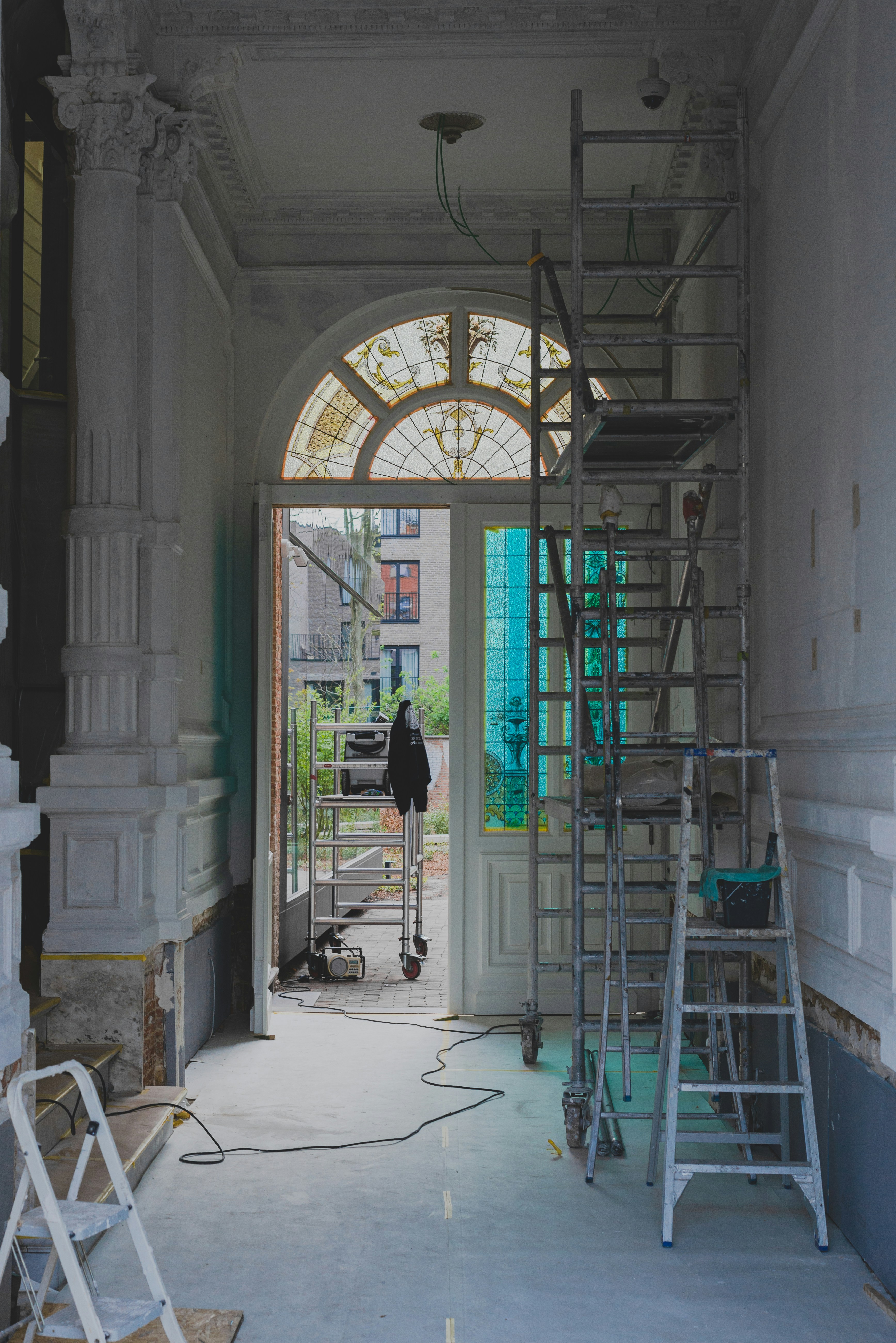 The image features a partially renovated interior space with ornate moldings and a large arched window with stained glass. Scaffolding and ladders are positioned throughout the area, along with painting supplies and construction equipment. The light coming through the window creates a contrast with the otherwise muted tones of the room.