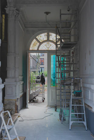 The image features a partially renovated interior space with ornate moldings and a large arched window with stained glass. Scaffolding and ladders are positioned throughout the area, along with painting supplies and construction equipment. The light coming through the window creates a contrast with the otherwise muted tones of the room.