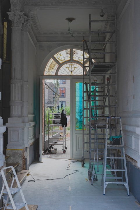 The image features a partially renovated interior space with ornate moldings and a large arched window with stained glass. Scaffolding and ladders are positioned throughout the area, along with painting supplies and construction equipment. The light coming through the window creates a contrast with the otherwise muted tones of the room.