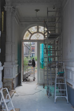 The image features a partially renovated interior space with ornate moldings and a large arched window with stained glass. Scaffolding and ladders are positioned throughout the area, along with painting supplies and construction equipment. The light coming through the window creates a contrast with the otherwise muted tones of the room.