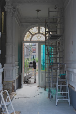 The image features a partially renovated interior space with ornate moldings and a large arched window with stained glass. Scaffolding and ladders are positioned throughout the area, along with painting supplies and construction equipment. The light coming through the window creates a contrast with the otherwise muted tones of the room.